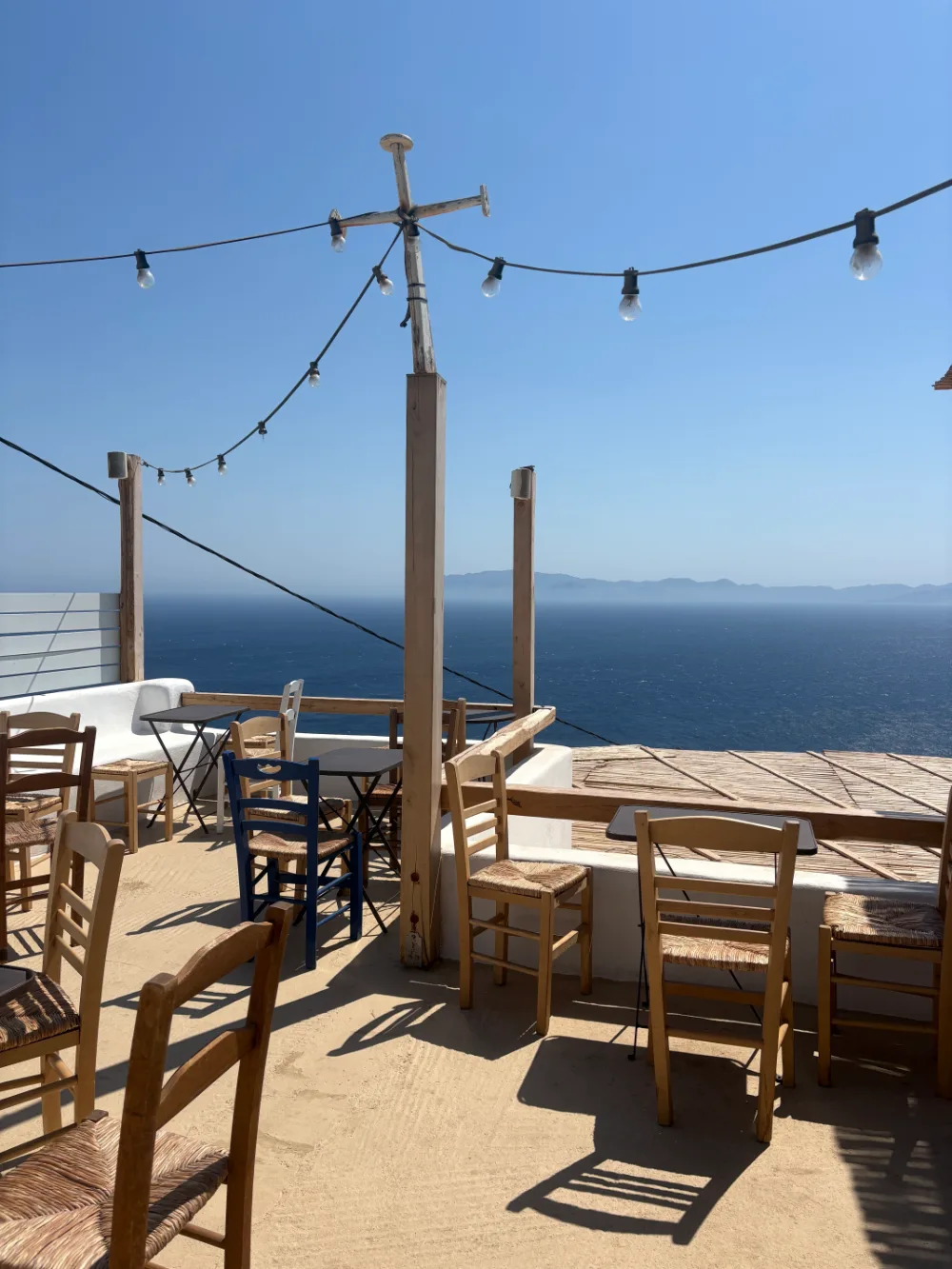 A taverna terrace above the sea in the Cyclades: rush-seated
                  chairs on a swept floor, string lights on a mast, and an
                  empty horizon.